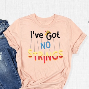 May include: A pink t-shirt with the text "I've Got No Strings" in colorful letters hanging from strings. The shirt is laid out on a white surface with a pair of blue jeans.