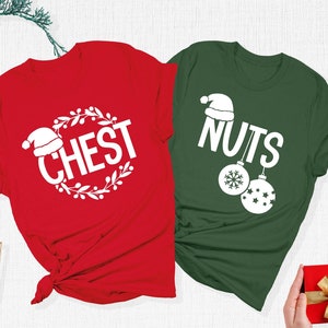 May include: Two Christmas-themed t-shirts, one red and one green, with white text and graphics. The red shirt says "CHEST" inside a wreath, and the green shirt says "NUTS" with ornament graphics. Festive holiday apparel.