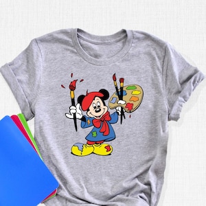 May include: A gray t-shirt with a cartoon image of Mickey Mouse dressed as an artist. He is holding paintbrushes and a paint palette. The shirt is a great gift for any Disney fan.