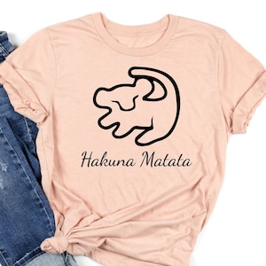 May include: A peach-colored t-shirt with a black outline of a lion's head and the text "Hakuna Matata". The t-shirt is paired with blue jeans.