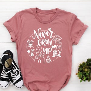 May include: A pink t-shirt with a white circle graphic that says "Never Grow Up" and includes various Disney park icons.