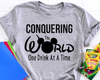 May include: A heather gray t-shirt with the text "CONQUERING the WORLD One Drink At A Time" in black. The word "World" features a Mickey Mouse head. Crayola crayons and colorful folders are also in the image.