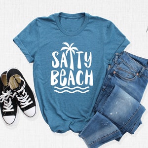 May include: A teal blue t-shirt with a white graphic that says "SAPPY BEACH" with a palm tree and waves. The shirt is laid out on a white surface with a pair of black sneakers and blue jeans.