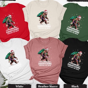 May include: A collection of t-shirts in various colors, including red, white, and black. Each shirt features a graphic of a Bigfoot wearing a Santa hat and carrying a Christmas tree, with the text "Squatching Through The Snow".