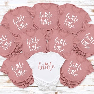 May include: Eight pink t-shirts with the text "bride tribe" and a white Mickey Mouse head design. One white t-shirt with the text "bride" and a white Mickey Mouse head design.