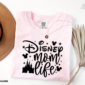 A pink t-shirt with the text "Disney mom life" printed on it, along with a castle silhouette and stars. The t-shirt is laid out on a white surface, and there is a brown hat and a palm frond to the side.
