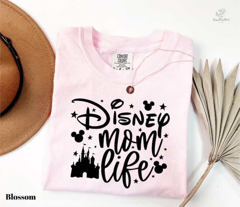 A pink t-shirt with the text "Disney mom life" printed on it, along with a castle silhouette and stars. The t-shirt is laid out on a white surface, and there is a brown hat and a palm frond to the side.