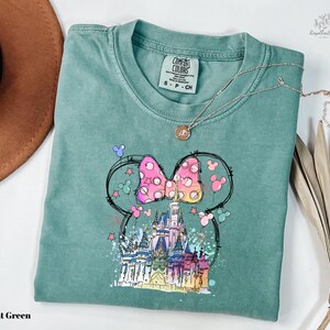 A light green t-shirt with a colorful illustration of a castle and Mickey Mouse characters on it, along with a necklace and a brown hat.