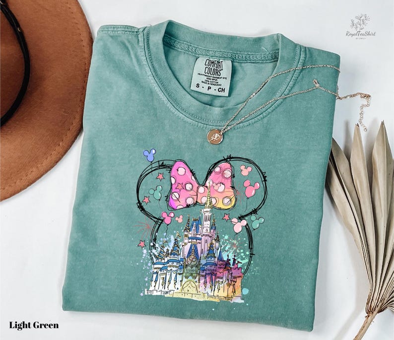 A light green t-shirt with a colorful illustration of a castle and Mickey Mouse characters on it, along with a necklace and a brown hat.