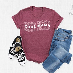 May include: A maroon t-shirt with the text "Cool Mama" repeated three times in white. The shirt is laid flat on a white surface with a pair of black sneakers and blue jeans.
