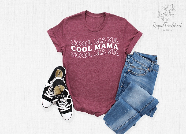 May include: A maroon t-shirt with the text "Cool Mama" repeated three times in white. The shirt is laid flat on a white surface with a pair of black sneakers and blue jeans.