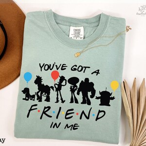 A light green t-shirt with a graphic design featuring silhouettes of characters from the animated film Toy Story, along with the text "You've Got a Friend in Me". The t-shirt is laid out on a white surface, accompanied by a hat, a necklace, and some decorative items.