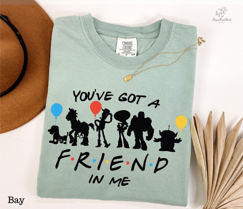 A light green t-shirt with a graphic design featuring silhouettes of characters from the animated film Toy Story, along with the text "You've Got a Friend in Me". The t-shirt is laid out on a white surface, accompanied by a hat, a necklace, and some decorative items.