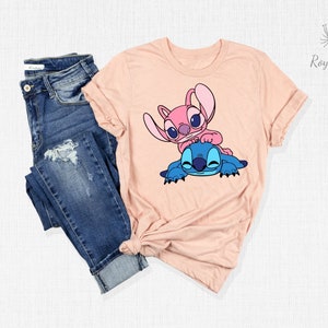 K&ouml;nnte beinhalten: Ein pfirsichfarbenes T-Shirt mit einem Motiv von Stitch und Angel aus dem Disney-Film Lilo & Stitch. Stitch ist blau und Angel ist rosa. Beide l&auml;cheln und umarmen sich.