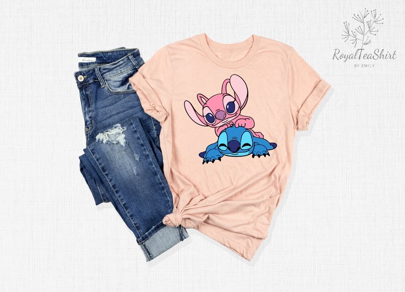 K&ouml;nnte beinhalten: Ein pfirsichfarbenes T-Shirt mit einem Motiv von Stitch und Angel aus dem Disney-Film Lilo & Stitch. Stitch ist blau und Angel ist rosa. Beide l&auml;cheln und umarmen sich.