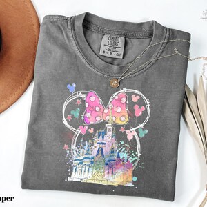 A gray t-shirt with a colorful graphic of a castle and Mickey Mouse characters printed on it, along with a necklace and a hat.