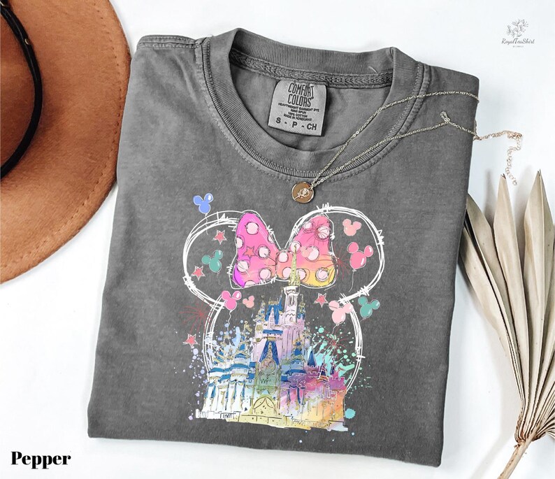 A gray t-shirt with a colorful graphic of a castle and Mickey Mouse characters printed on it, along with a necklace and a hat.