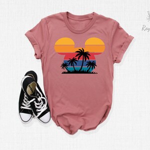 May include: A pink t-shirt with a retro sunset and palm trees in the shape of Mickey Mouse's head.