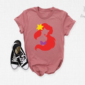 May include: A pink t-shirt with a red silhouette of Ariel, a Disney princess, with a yellow star on her head. The silhouette is in a simple, minimalist style.