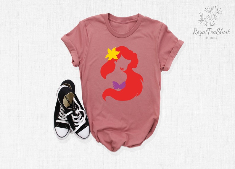 May include: A pink t-shirt with a red silhouette of Ariel, a Disney princess, with a yellow star on her head. The silhouette is in a simple, minimalist style.