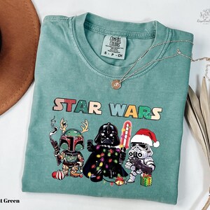 A light green t-shirt with a Star Wars graphic design featuring three characters: a knight, a cowboy, and a cowboy hat. The t-shirt is laid out on a white surface, with a brown hat and some dried leaves visible in the background.