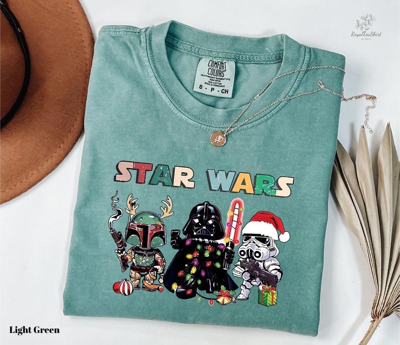 A light green t-shirt with a Star Wars graphic design featuring three characters: a knight, a cowboy, and a cowboy hat. The t-shirt is laid out on a white surface, with a brown hat and some dried leaves visible in the background.