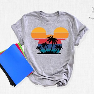 May include: Gray t-shirt with a retro sunset design in the shape of Mickey Mouse ears. The sunset has orange, yellow, red, blue, and green stripes. There are palm trees in front of the sunset.