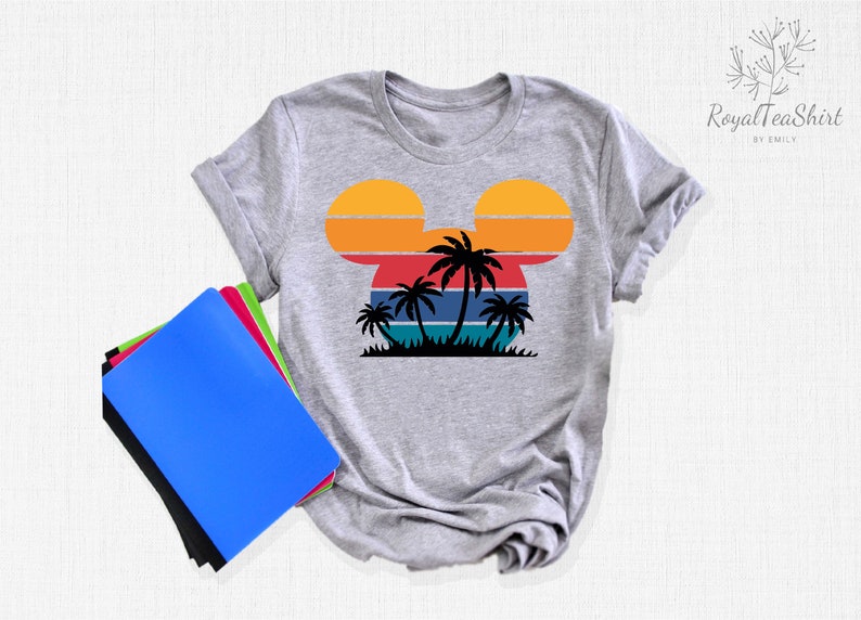 May include: Gray t-shirt with a retro sunset design in the shape of Mickey Mouse ears. The sunset has orange, yellow, red, blue, and green stripes. There are palm trees in front of the sunset.
