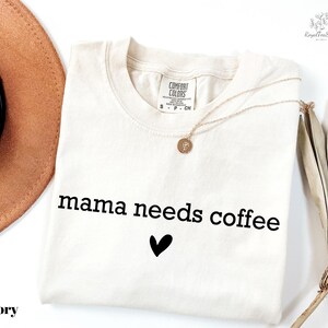 A white t-shirt with the text "mama needs coffee" printed on it, along with a heart symbol. The t-shirt is folded and placed on a white surface, with a brown hat and a plant in the background.