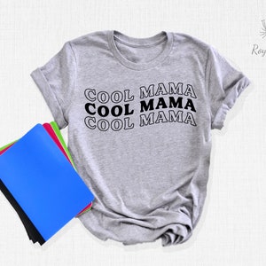 May include: A gray t-shirt with the text "COOL MAMA" repeated three times in black with a white outline. The shirt is folded and lying on a white surface with a stack of notebooks.