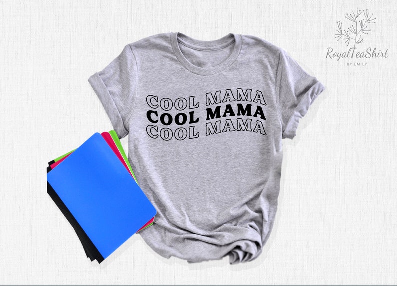 May include: A gray t-shirt with the text "COOL MAMA" repeated three times in black with a white outline. The shirt is folded and lying on a white surface with a stack of notebooks.