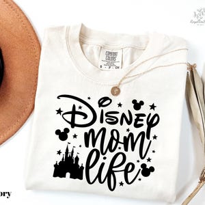 A white t-shirt with a black graphic design that says "Disney mom life" and features a castle silhouette and stars. The t-shirt is laid out on a white background, along with a brown hat and a fan.