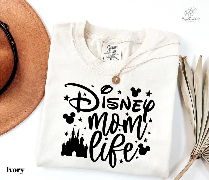 A white t-shirt with a black graphic design that says "Disney mom life" and features a castle silhouette and stars. The t-shirt is laid out on a white background, along with a brown hat and a fan.