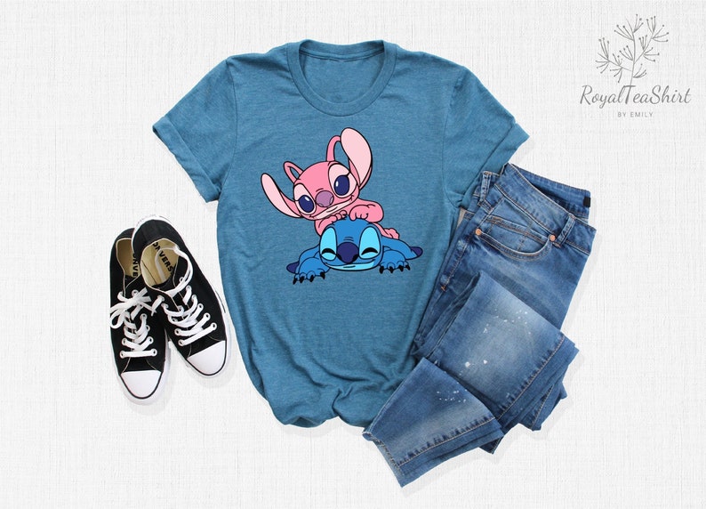K&ouml;nnte beinhalten: Ein t&uuml;rkisfarbenes T-Shirt mit einem pinkfarbenen und blauen Stitch-Charakter-Motiv. Das Shirt liegt auf einer wei&szlig;en Oberfl&auml;che mit einem Paar schwarzer Turnschuhe und einer blauen Jeans.