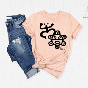 May include: A pink t-shirt with a black design of a frog and a sun with a face. The text "Taino" is below the design.