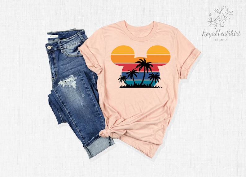 May include: A pink t-shirt with a retro sunset design in the shape of Mickey Mouse's head. Palm trees are silhouetted in front of the sunset. The shirt is paired with blue jeans.