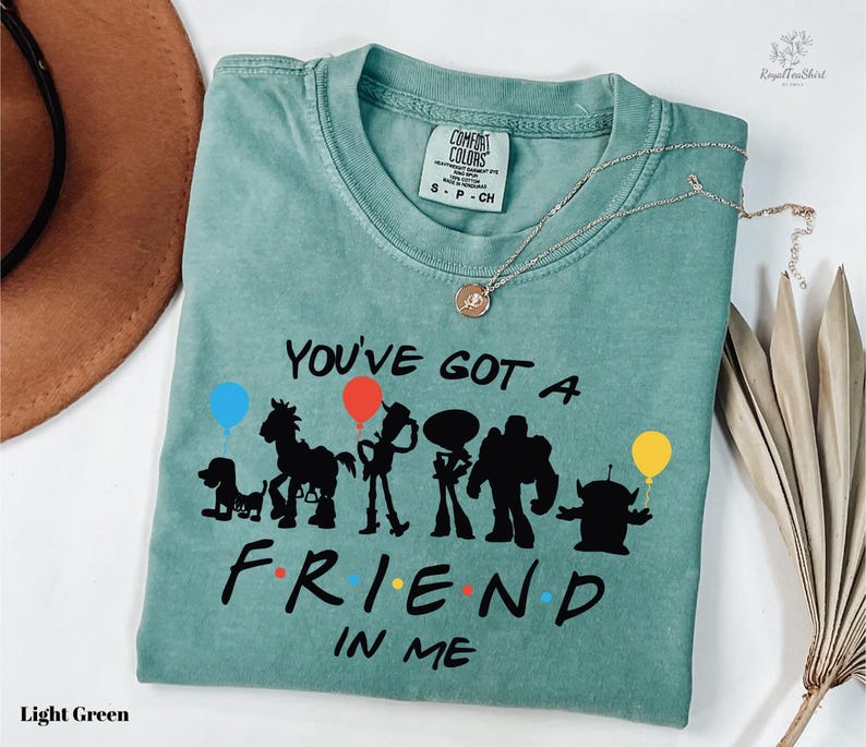 A light green t-shirt with a graphic design featuring silhouettes of characters from the animated film Toy Story, along with the text "You've got a friend in me" printed on it.