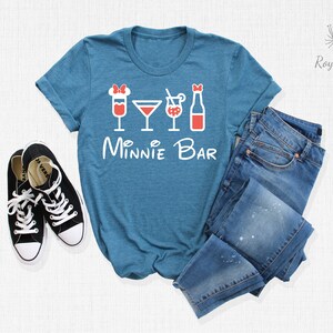 May include: A teal blue t-shirt with a white graphic of four drinks and the text "Minnie Bar". The t-shirt is laid out on a white surface with a pair of black sneakers and a pair of blue jeans.