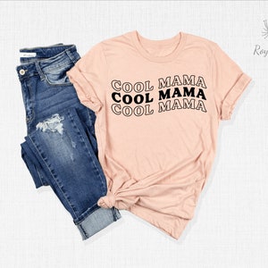 May include: A pink t-shirt with the text "COOL MAMA" repeated three times in black, stacked vertically. The shirt is shown on a white background with a pair of blue jeans.