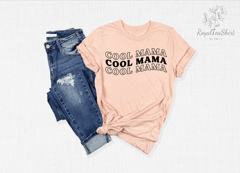 May include: A pink t-shirt with the text "COOL MAMA" repeated three times in black, stacked vertically. The shirt is shown on a white background with a pair of blue jeans.