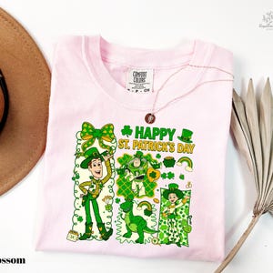 A pink t-shirt with a cartoon design of St. Patrick's Day characters, including a man, a woman, and a dog, along with the text "Happy St. Patrick's Day". The t-shirt is displayed on a white background, accompanied by a brown hat and a palm frond.
