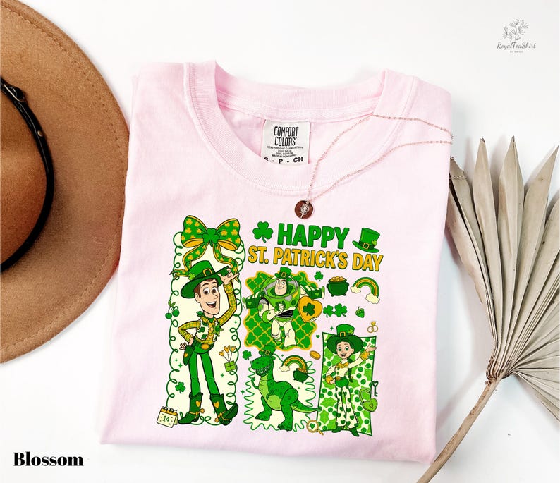 A pink t-shirt with a cartoon design of St. Patrick's Day characters, including a man, a woman, and a dog, along with the text "Happy St. Patrick's Day". The t-shirt is displayed on a white background, accompanied by a brown hat and a palm frond.
