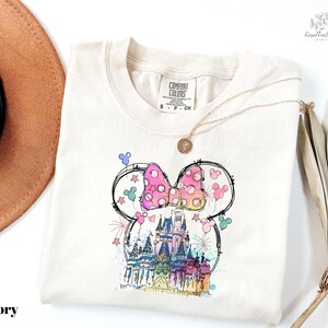 A white t-shirt with a colorful illustration of a castle and Mickey Mouse characters printed on it, along with a necklace and a hat.