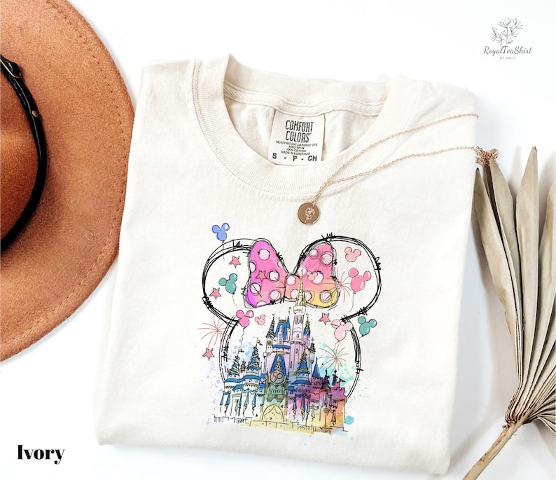 A white t-shirt with a colorful illustration of a castle and Mickey Mouse characters printed on it, along with a necklace and a hat.