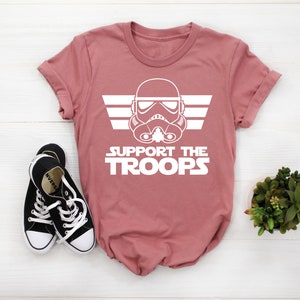 May include: A pink t-shirt with a white graphic of a Star Wars Stormtrooper helmet with wings and the text "SUPPORT THE TROOPS".