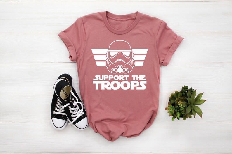 May include: A pink t-shirt with a white graphic of a Star Wars Stormtrooper helmet with wings and the text "SUPPORT THE TROOPS".