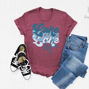 May include: A maroon t-shirt with the text "Lake Life" in blue and white lettering. The shirt is laid flat on a white surface with a pair of black sneakers and blue jeans.