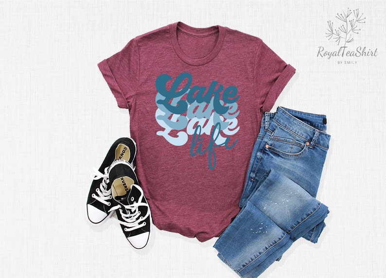 May include: A maroon t-shirt with the text "Lake Life" in blue and white lettering. The shirt is laid flat on a white surface with a pair of black sneakers and blue jeans.
