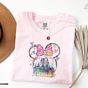 A pink t-shirt with a whimsical Mickey Mouse design, including a castle and fireworks, is displayed alongside a brown hat and a palm frond.