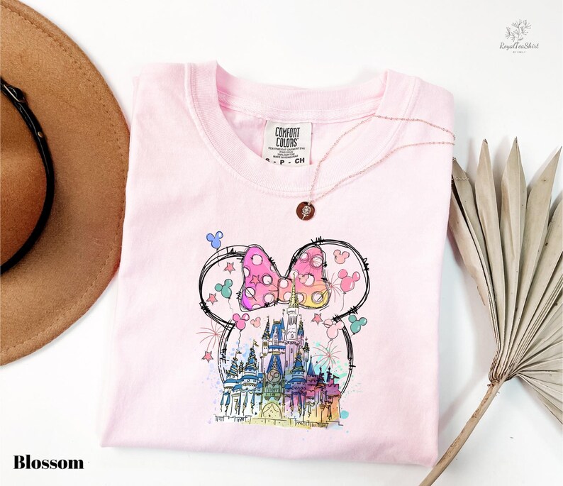 A pink t-shirt with a whimsical Mickey Mouse design, including a castle and fireworks, is displayed alongside a brown hat and a palm frond.